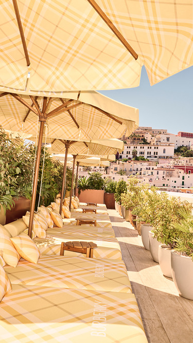 Outdoor lounge area with yellow plaid umbrellas and cushions, surrounded by potted plants, overlooking a hillside town.