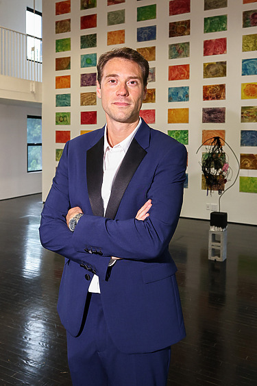 Man in a blue suit standing in an art gallery with colorful abstract paintings on the wall behind him.