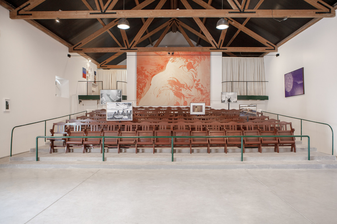 Interior of an empty hall with rows of wooden chairs facing a large abstract mural and several framed artworks.