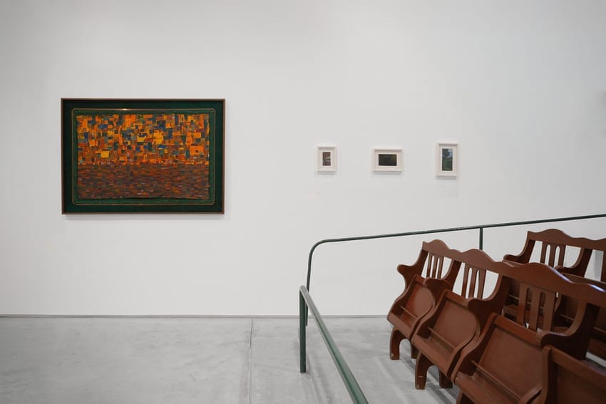 Art gallery with a large colorful abstract painting and three smaller framed artworks on a white wall, near wooden chairs.