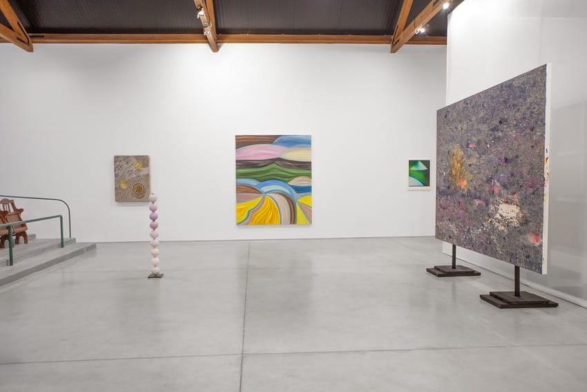 Contemporary art gallery interior with abstract paintings on white walls and a vertical sculpture in the center.