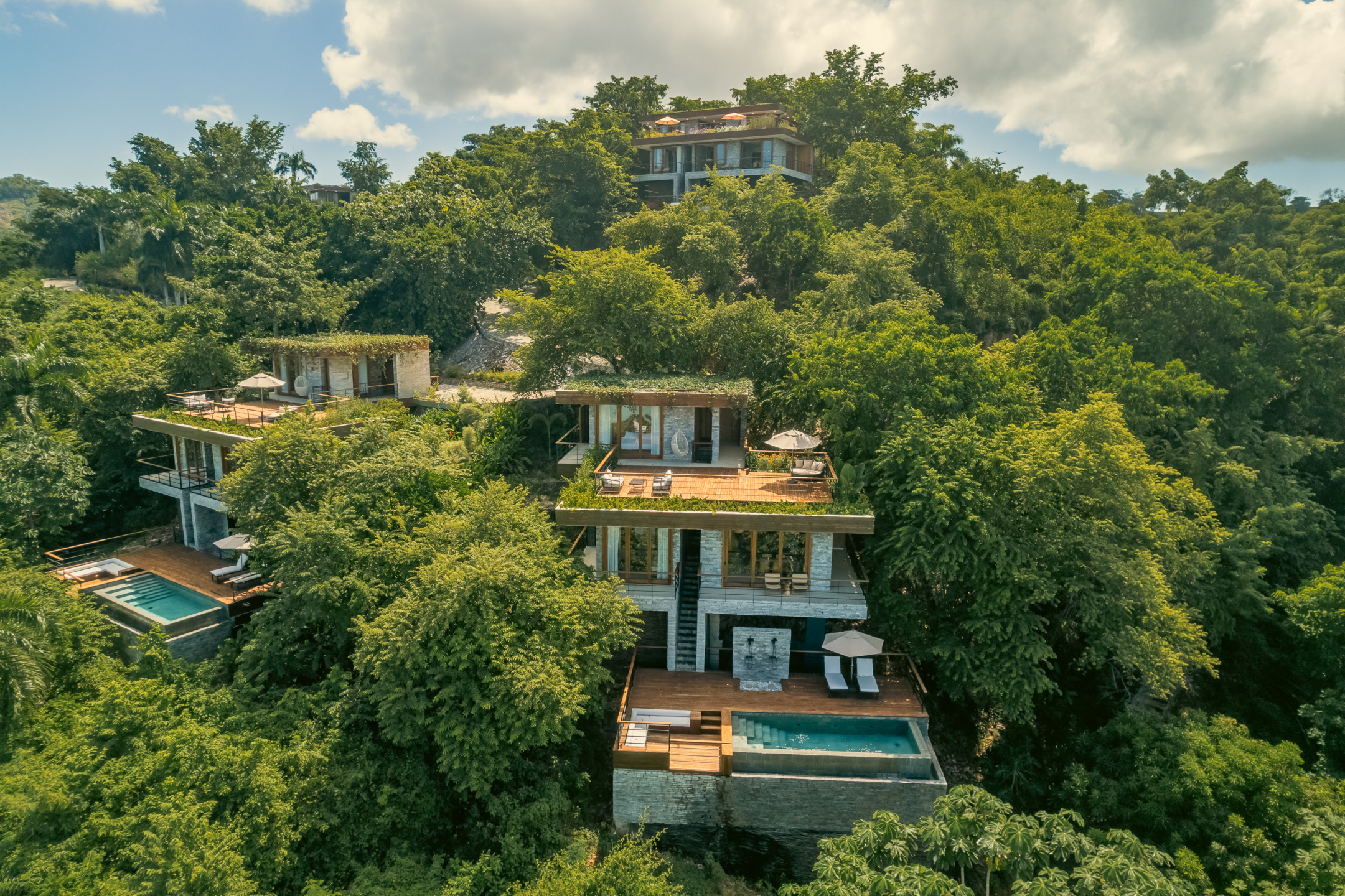 Luxurious hillside villas surrounded by lush greenery and private pools under a sunny sky.