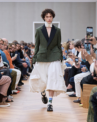 Model walking down a runway wearing a green jacket and wide white pants during a fashion show.