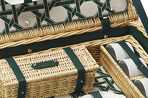 Close-up of an open wicker picnic basket with green straps, showing interior compartments and white cups.