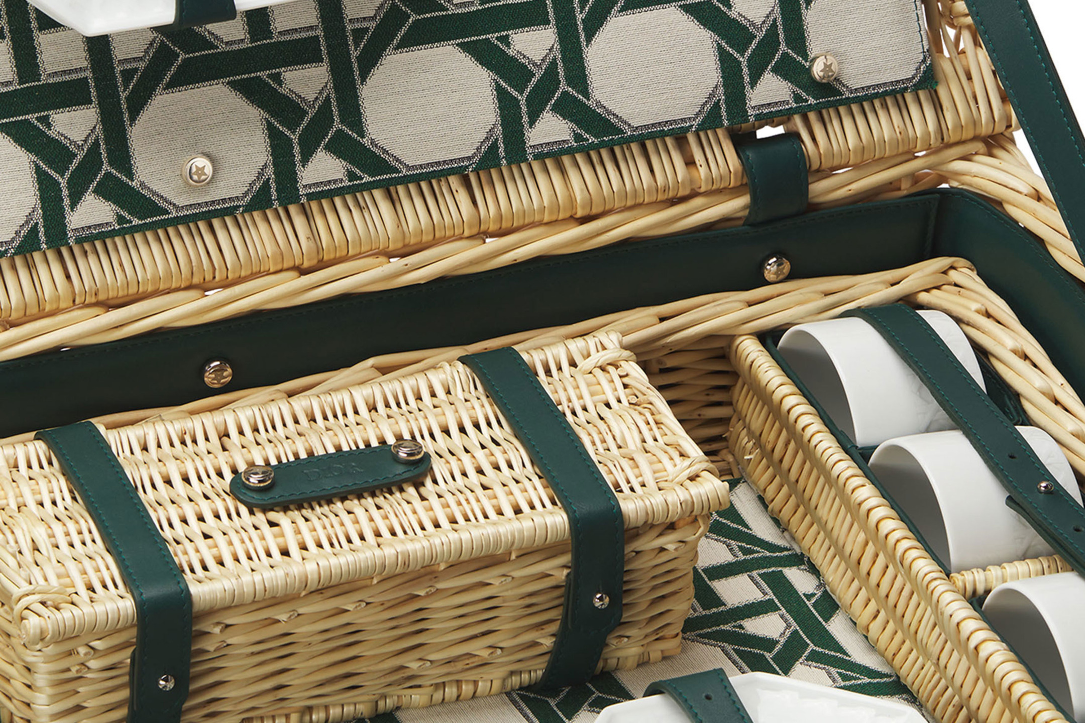 Close-up of an open wicker picnic basket with green straps, showing interior compartments and white cups.