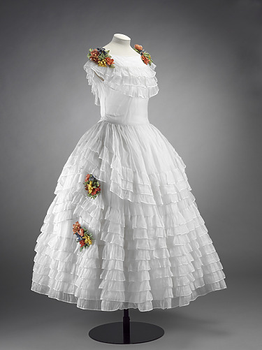 White ruffled dress with colorful floral accents displayed on a mannequin against a gray background.