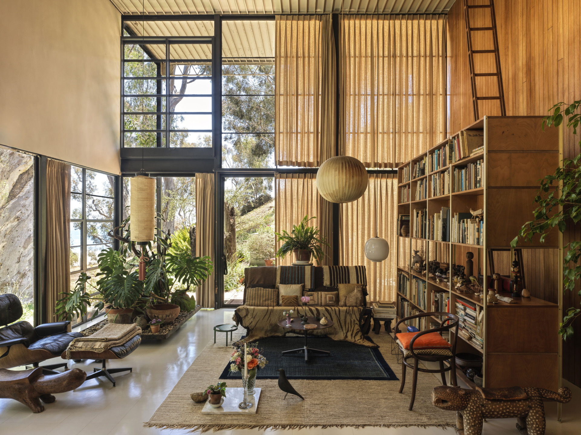 Mid-century modern living room with large windows, tropical plants, wooden shelves, and cozy seating.