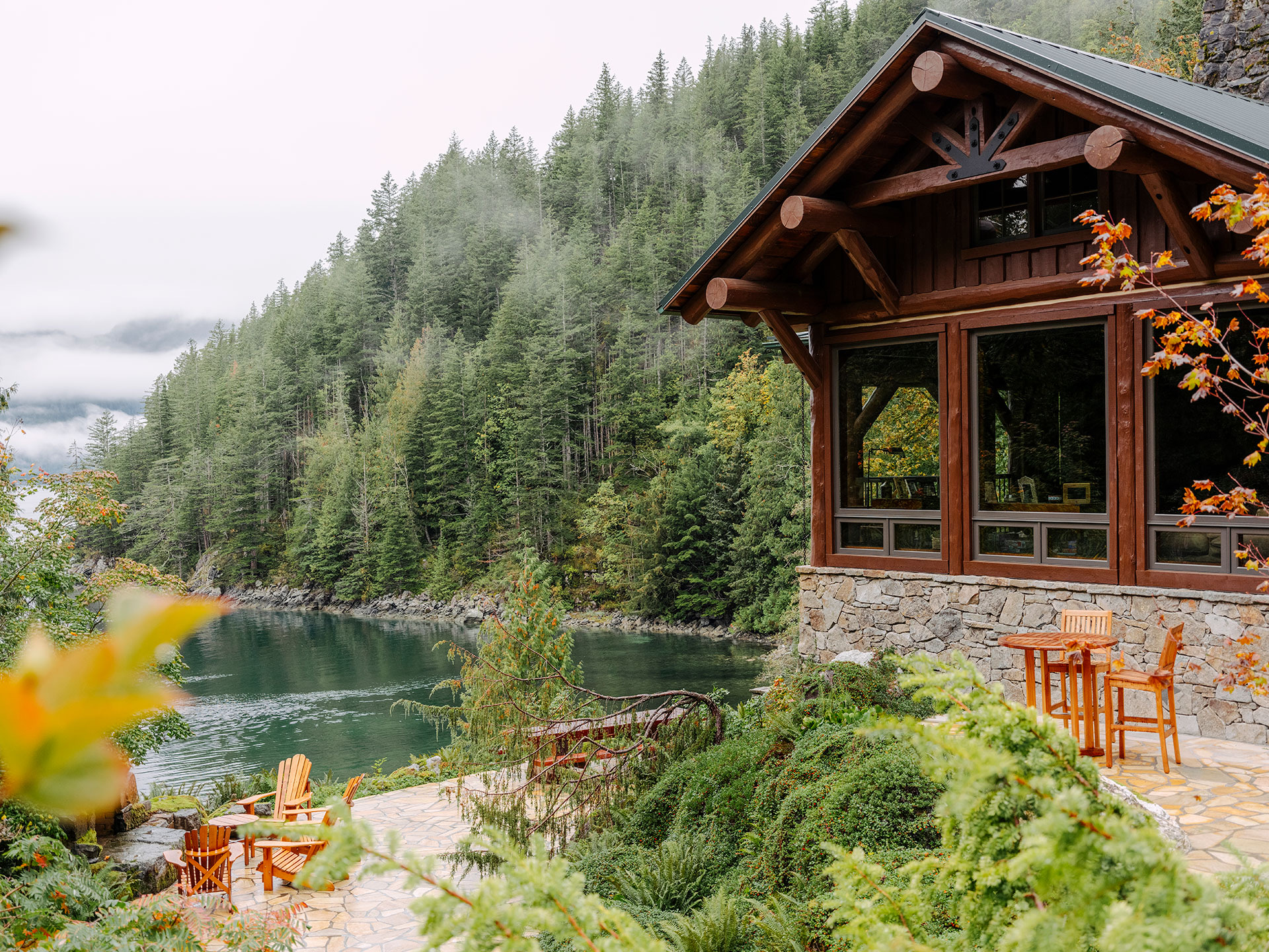 Lakeside cabin with wooden chairs on a stone patio, surrounded by lush green forest and misty mountains in the background.