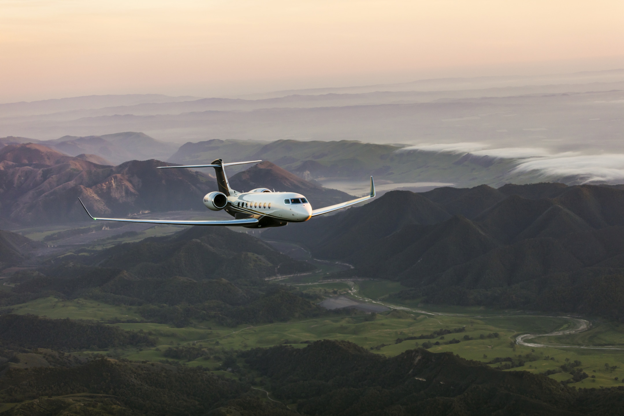 Private jet flying over lush green mountains during sunset, with a clear sky and distant rolling hills in the background.