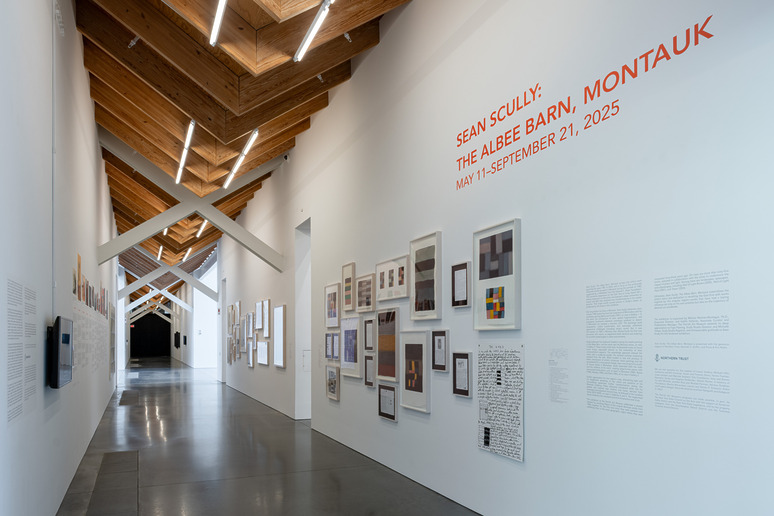 Art exhibition hallway with various framed paintings and text on white walls, wooden ceiling above, exhibition details in text.