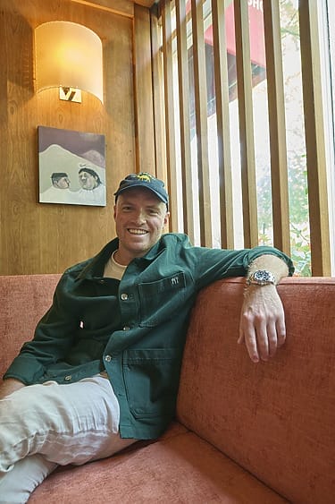 Man in green jacket and cap smiling while sitting on a red sofa in a cozy room with wooden walls and a small painting.