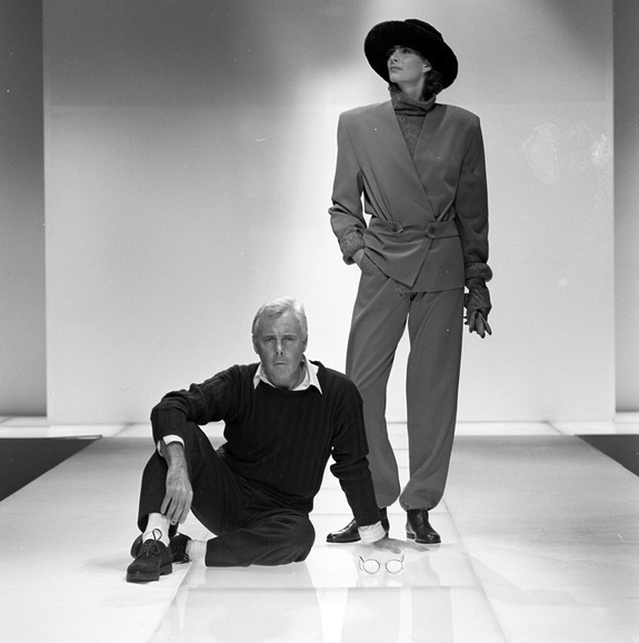 Fashion designer seated on runway beside model in stylish outfit with hat during a fashion show.