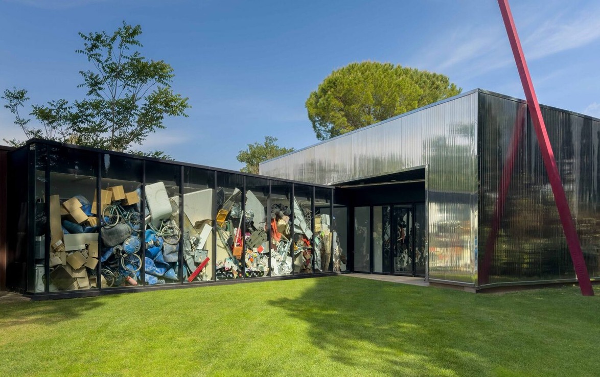 Modern building exterior with large windows filled with colorful recycling materials, surrounded by green lawn and trees.