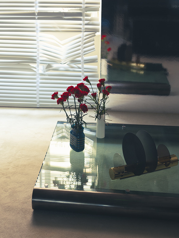 Modern living room with a glass table, red flowers in vases, and soft lighting from a window with horizontal blinds.