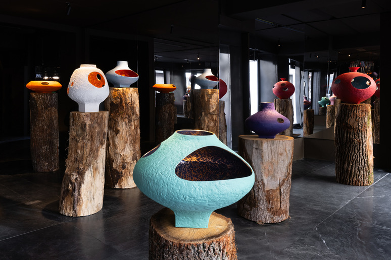 Colorful, abstract pottery displayed on wooden stumps in a dimly lit gallery environment with reflective surfaces.