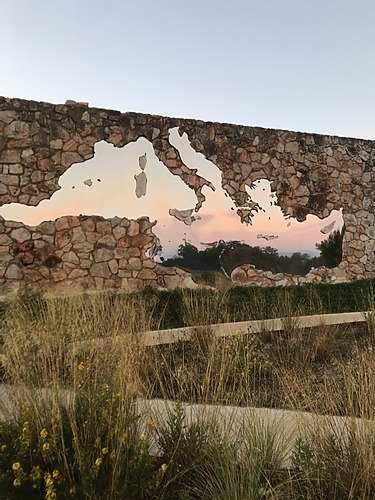 Stone wall with a mirrored map cutout reflecting a sunset sky, surrounded by dry grass and wildflowers in the foreground.