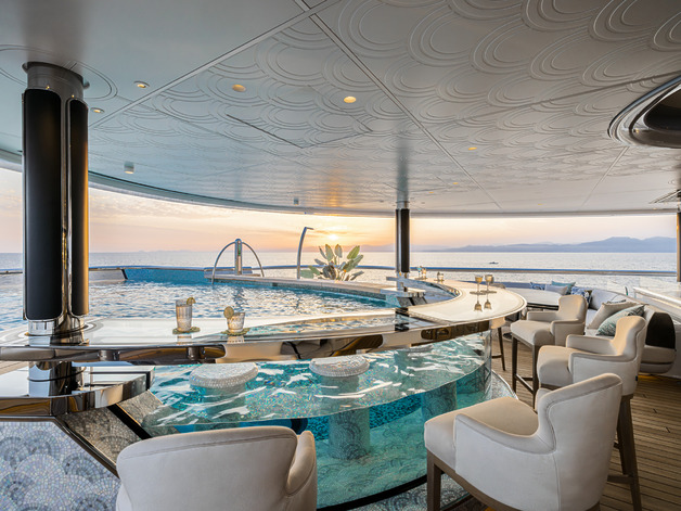 Luxurious yacht deck with circular pool, bar, and ocean view at sunset in the background.
