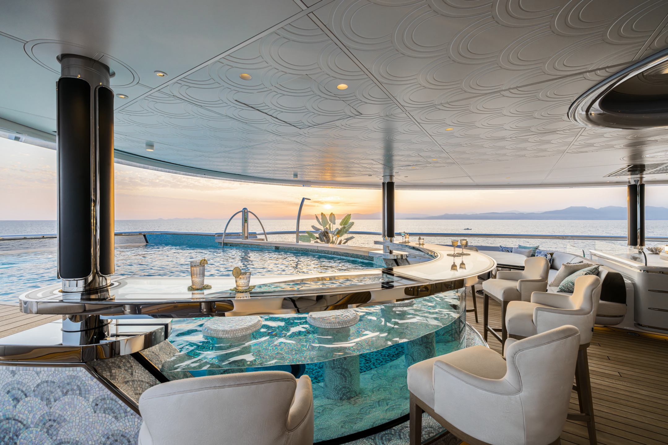 Luxurious yacht deck with circular pool, bar, and ocean view at sunset in the background.