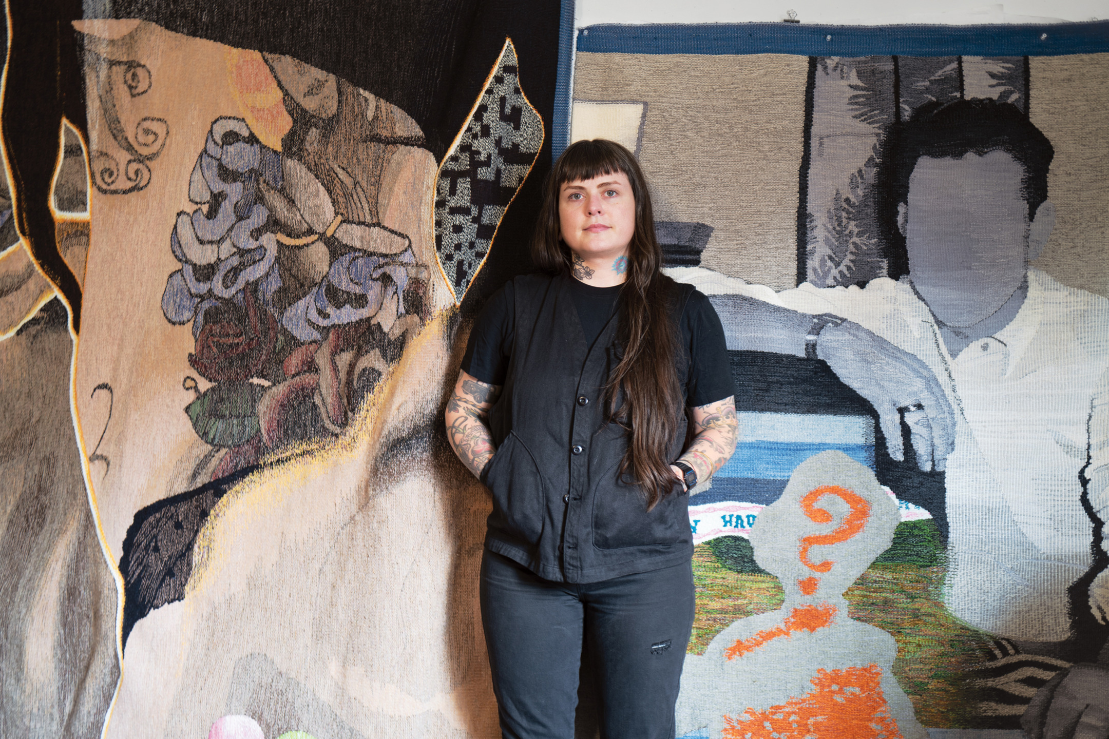 Person with tattoos standing between two large artistic tapestries in a gallery setting.
