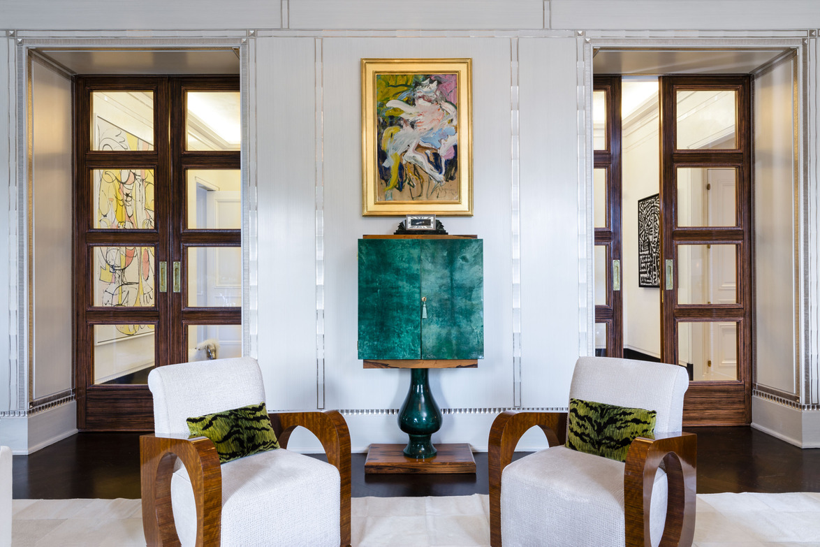 Elegant living room with two wooden armchairs, green cabinet, abstract art on walls, and natural light from large windows.