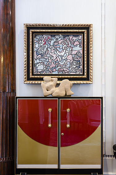 Abstract painting above cabinet with red and gold doors, adorned with a reclining stone sculpture.