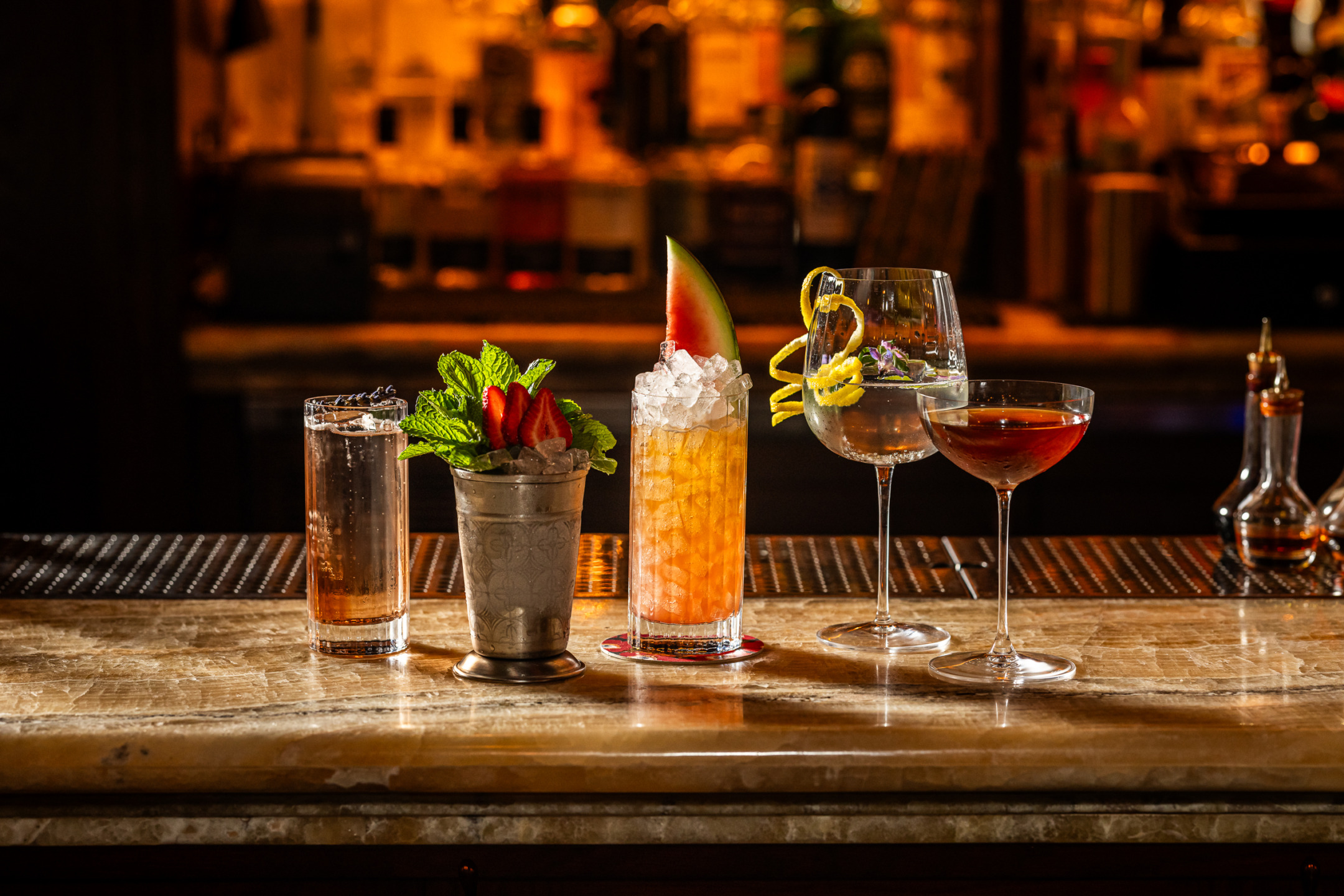 Assorted cocktails on a marble bar counter decorated with fruits and garnishes in a dimly lit bar setting.