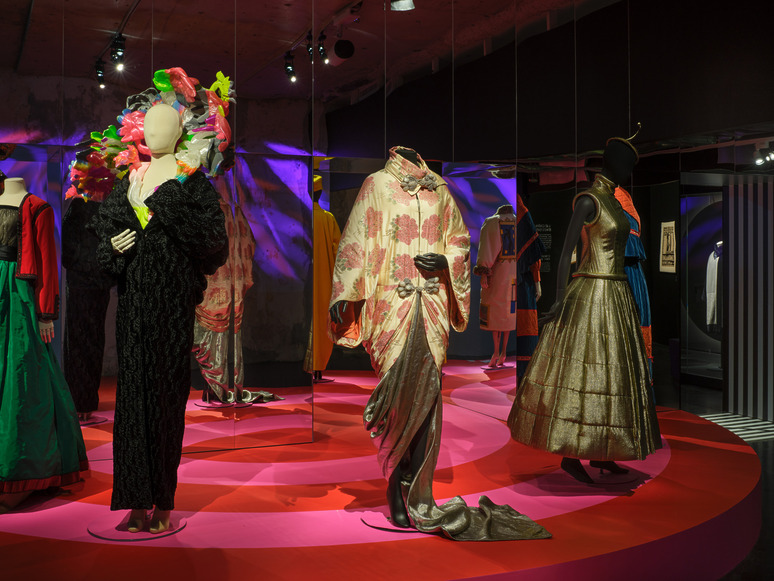 Fashion exhibit showcasing mannequins in elaborate colorful dresses on a vibrant circular pink platform in a dimly lit room.