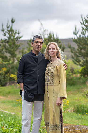 A man and a woman standing together outdoors in a lush green landscape with trees and hills in the background.