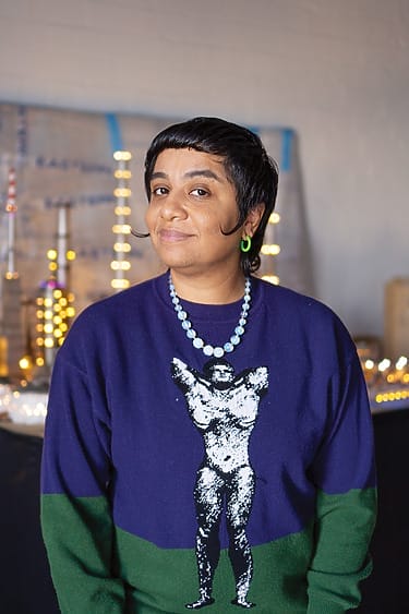 Person wearing a blue and green sweater with a graphic design, standing in a room with string lights in the background.