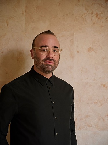 Person with glasses and beard wearing a black shirt against a textured beige wall.