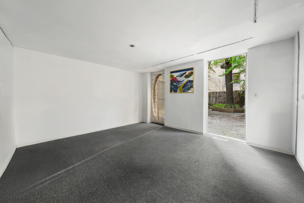 Sunny empty room with white walls, gray carpet, single wall art, open door leading to a garden with greenery.