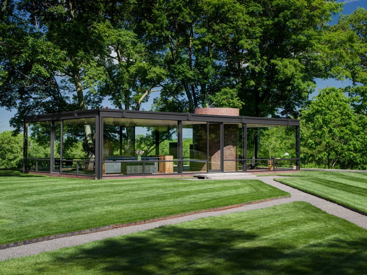 Modern glass house with steel frame set in a lush green landscape with neatly trimmed grass and large trees.