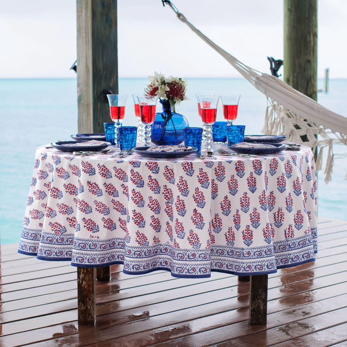 Round table with floral tablecloth, blue glassware, red drinks, and a flower centerpiece, set by the seaside with a hammock nearby.