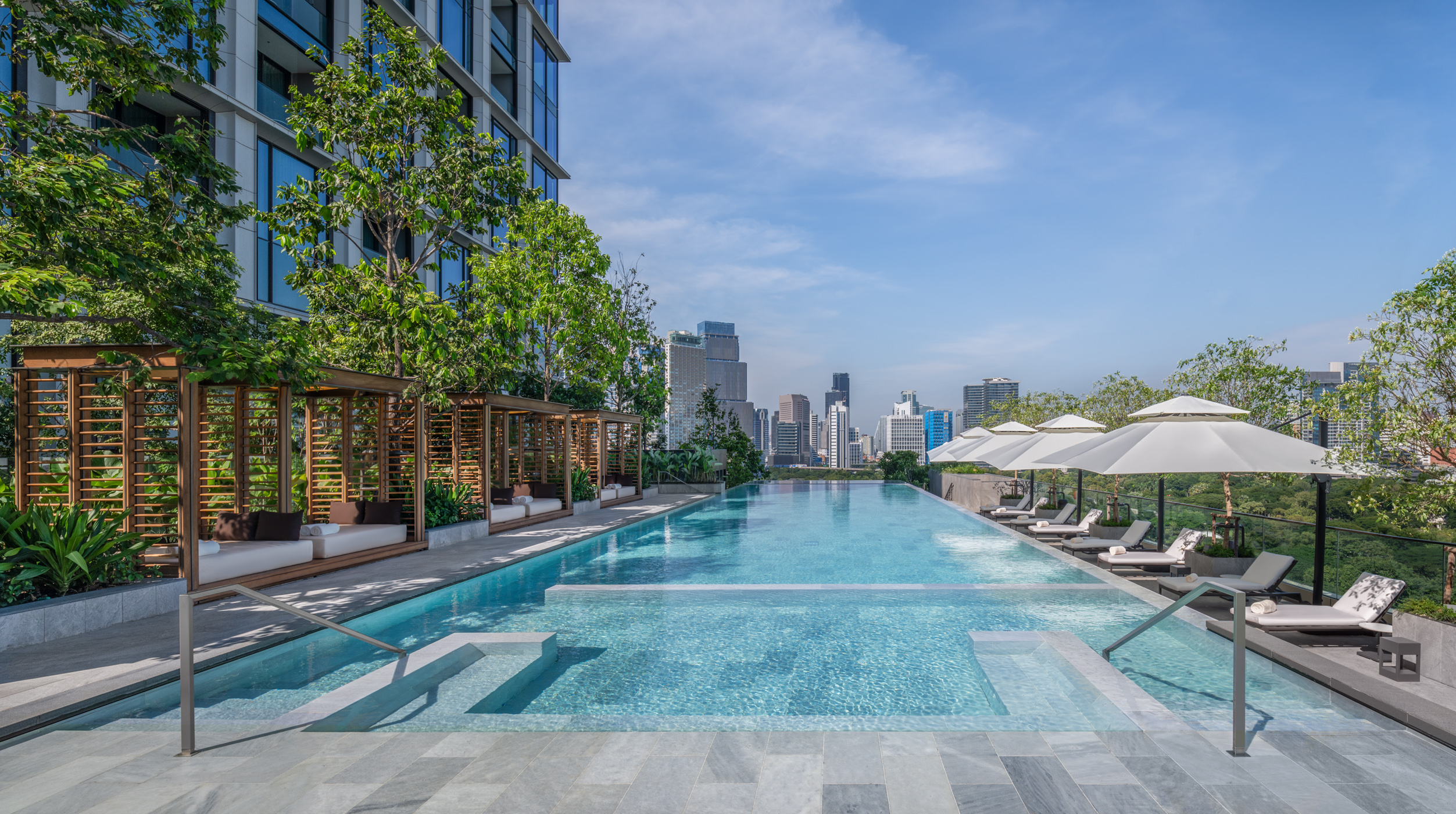 Rooftop infinity pool with lounge chairs and cabanas overlooking a city skyline on a clear, sunny day.