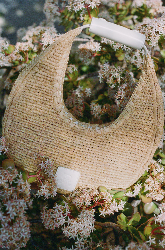 Woven crescent-shaped handbag with white handle resting on flowering shrub.