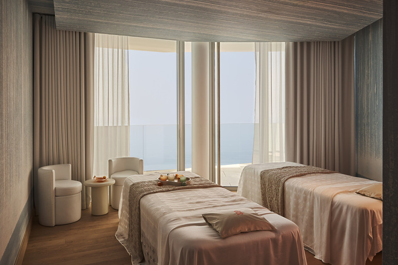 Luxurious spa room with two massage tables, soft lighting, sheer curtains, and ocean view balcony for relaxing ambiance.