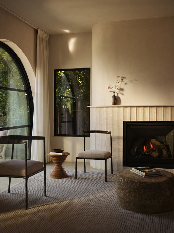 Cozy living room with armchairs, a small table, fireplace, vase with flowers, and large arched window with curtains.