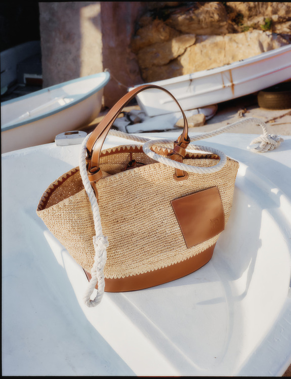 Woven straw handbag with leather accents resting in a small white boat, surrounded by ropes and nautical background.