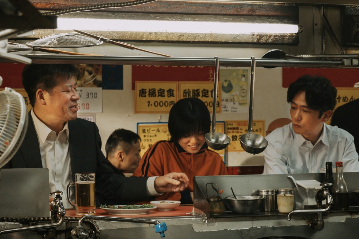 Three people sitting at a Japanese food stall, enjoying a meal and chatting over drinks and food on the counter.