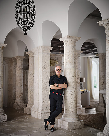 Man in black outfit leaning on stone column in elegant interior with arched doorways and large geometric pendant light overhead