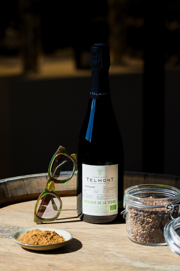 Bottle of Telmont champagne on a wooden table with sunglasses and small jars of spices in sunlight.
