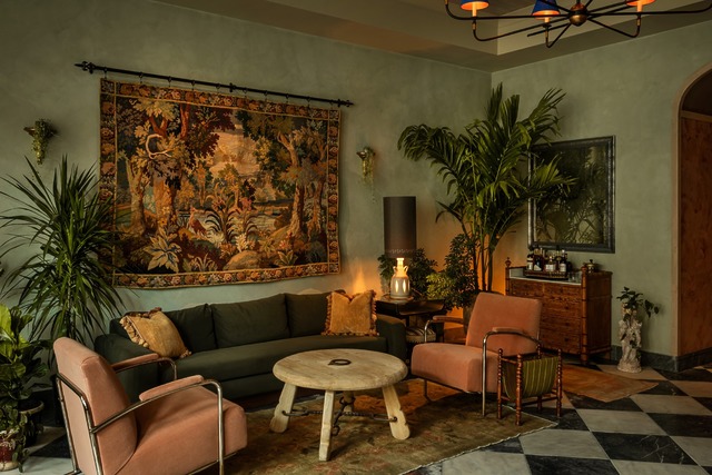 Cozy living room with green sofa, tapestry, plants, armchairs, wooden table, and chandelier for a warm, stylish ambiance.