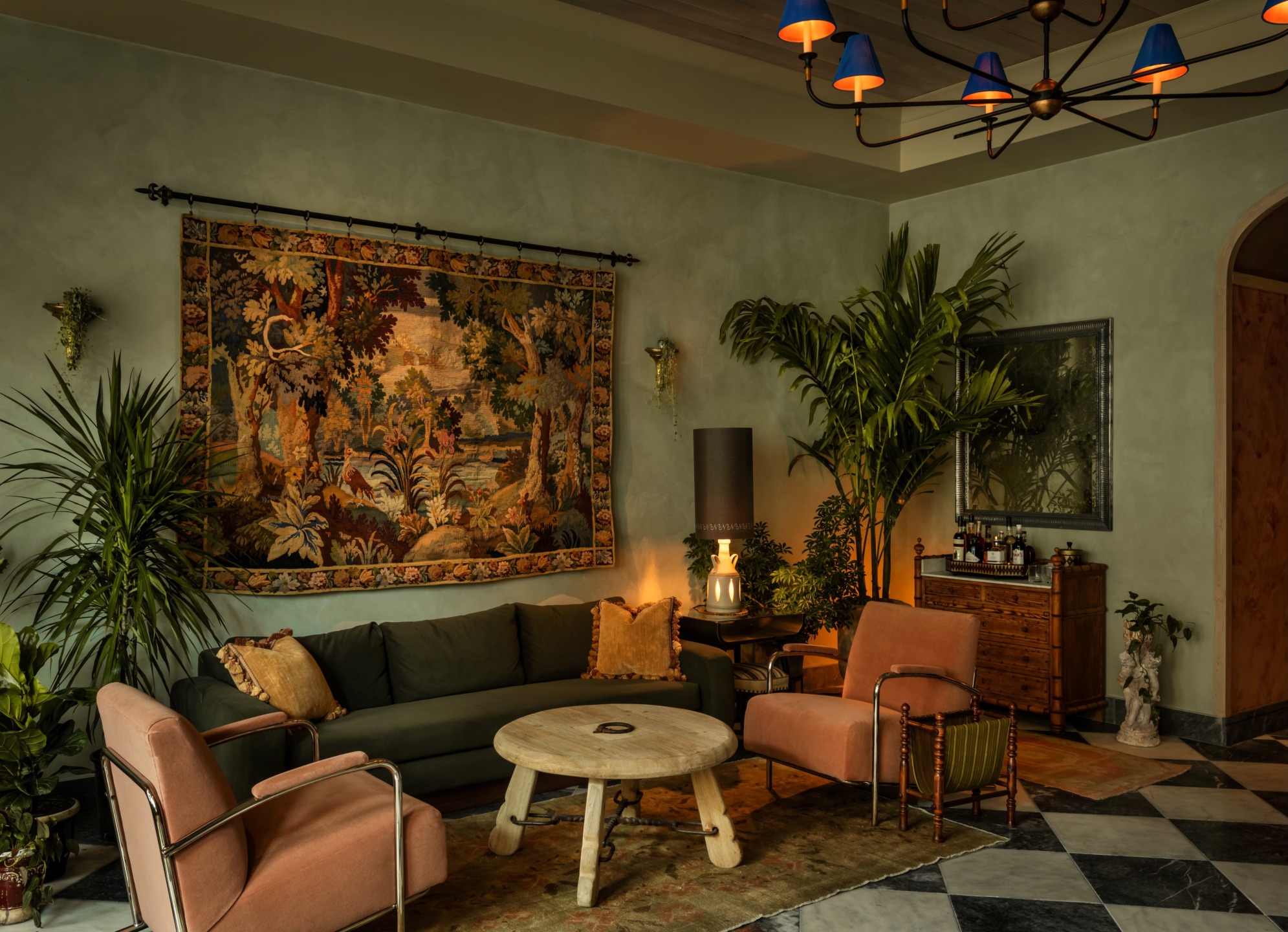 Cozy living room with green sofa, tapestry, plants, armchairs, wooden table, and chandelier for a warm, stylish ambiance.