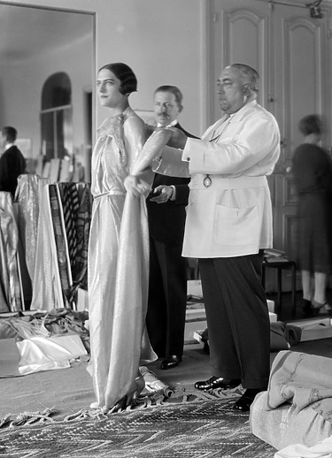 A designer fits a glamorous gown on a model in a fashion studio, surrounded by luxurious fabrics and attentive staff.