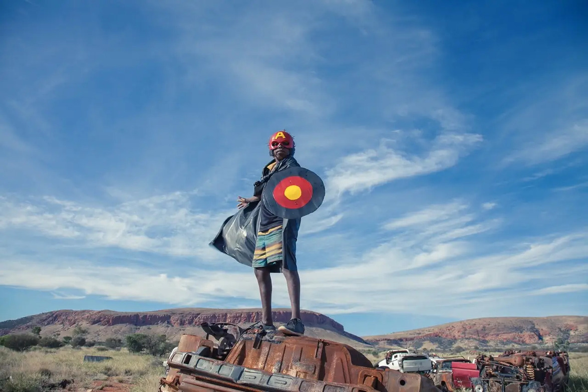 Person in colorful attire stands confidently on a rusty vehicle under a blue sky with clouds in a rural landscape.