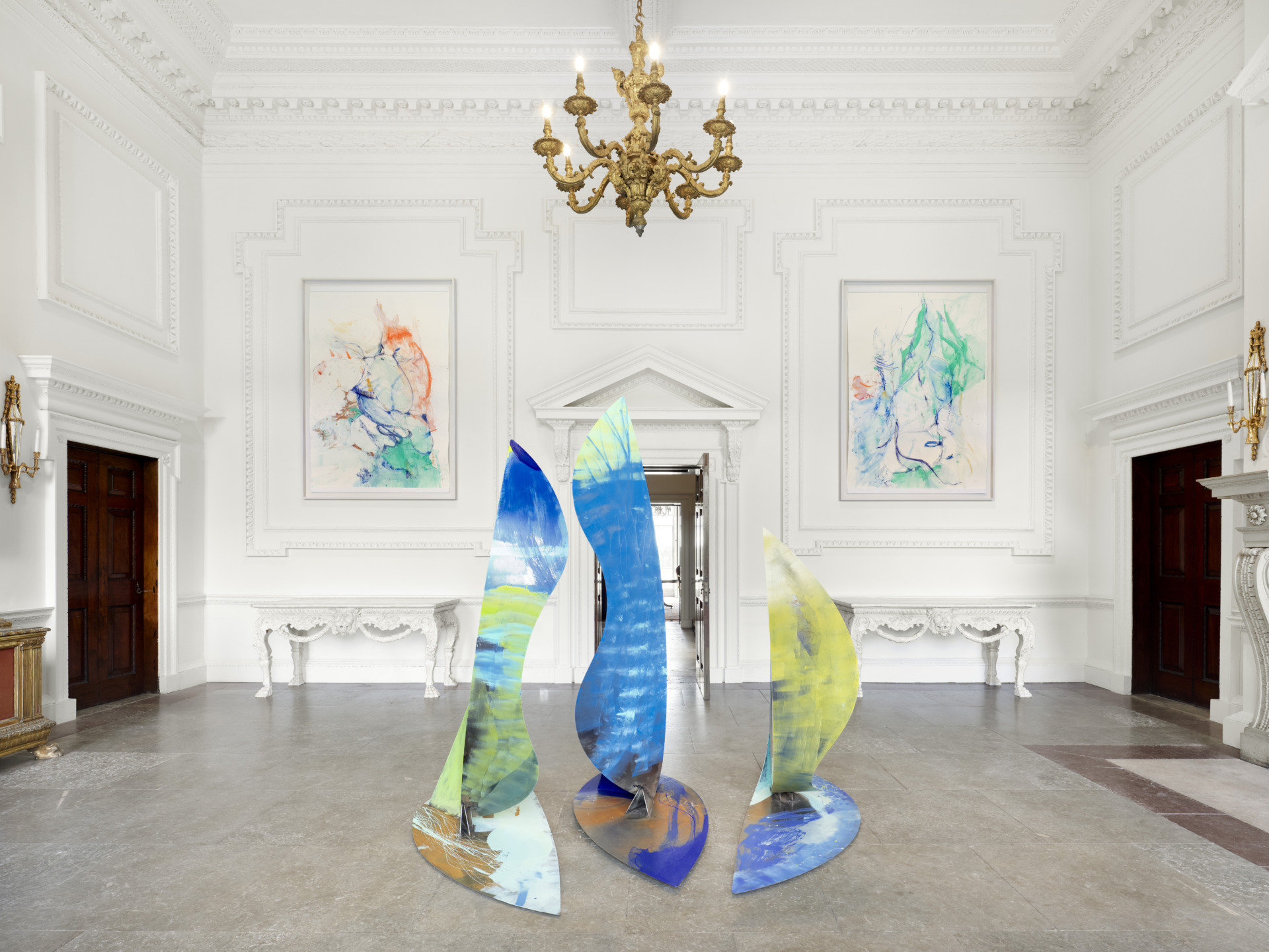Colorful abstract sculptures in an elegant room with white walls, artwork on display, and an ornate chandelier above.