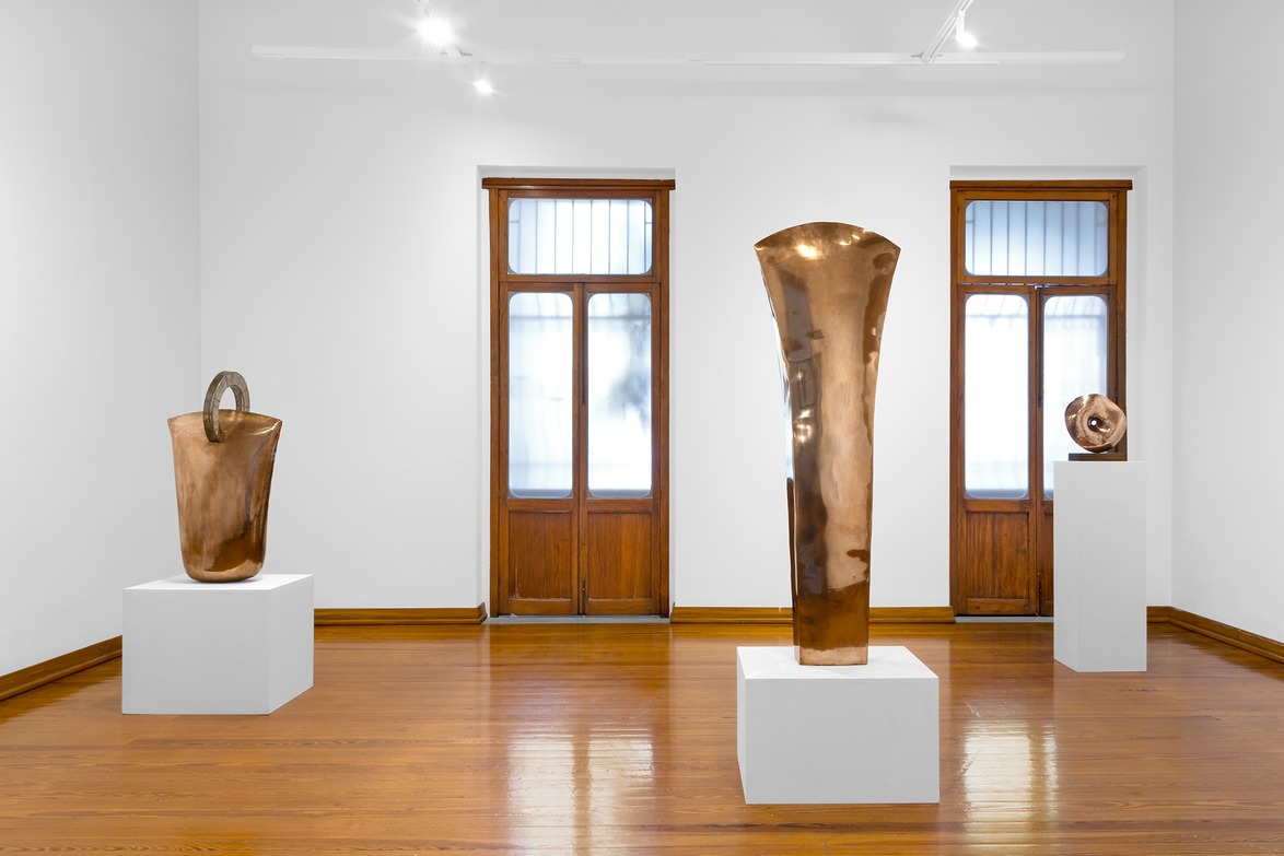 Contemporary art gallery displaying three abstract bronze sculptures on white pedestals in a room with wooden floors.