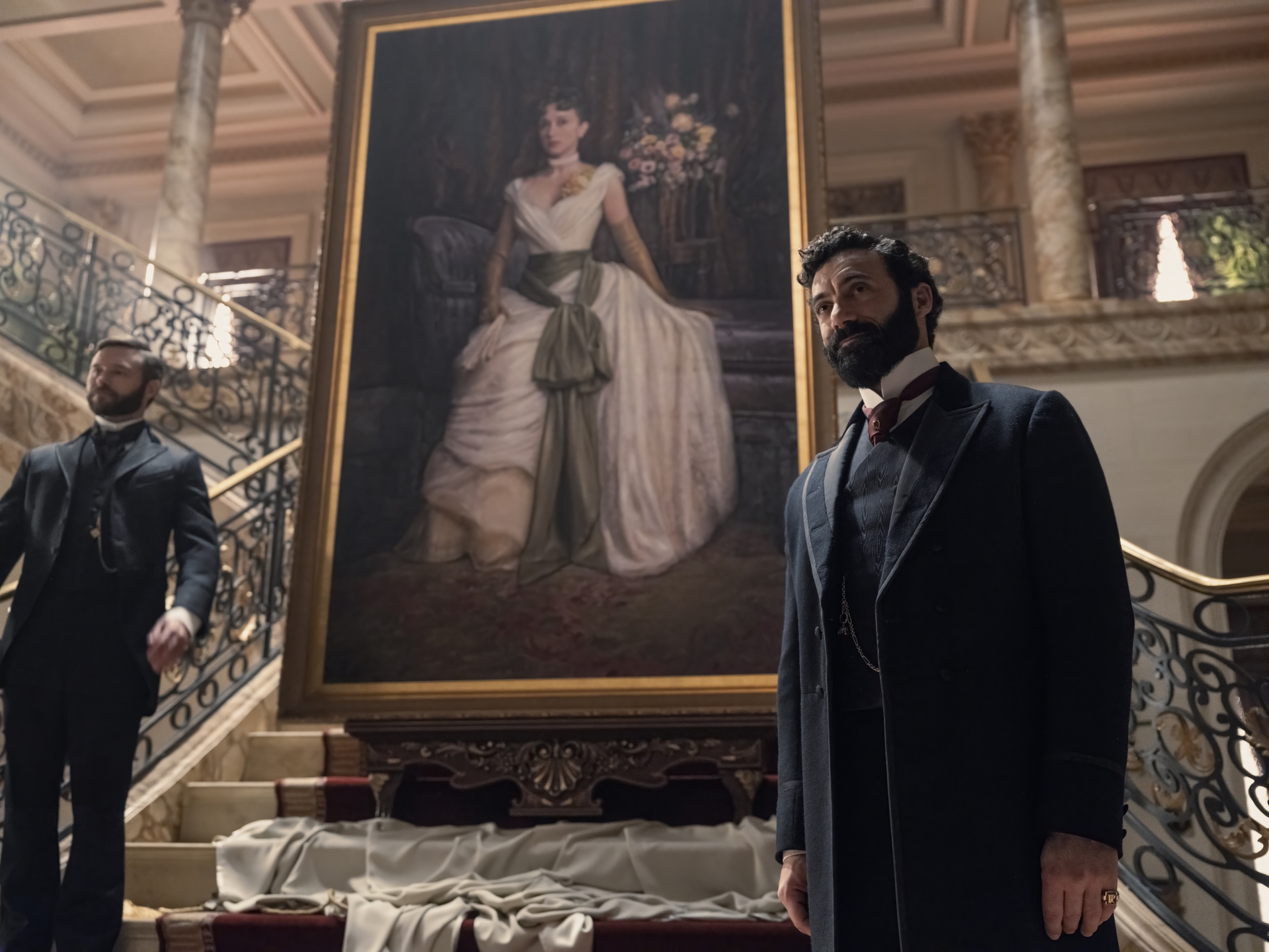 Two people standing near a grand staircase with a large portrait of a woman in an elegant gown in the background.