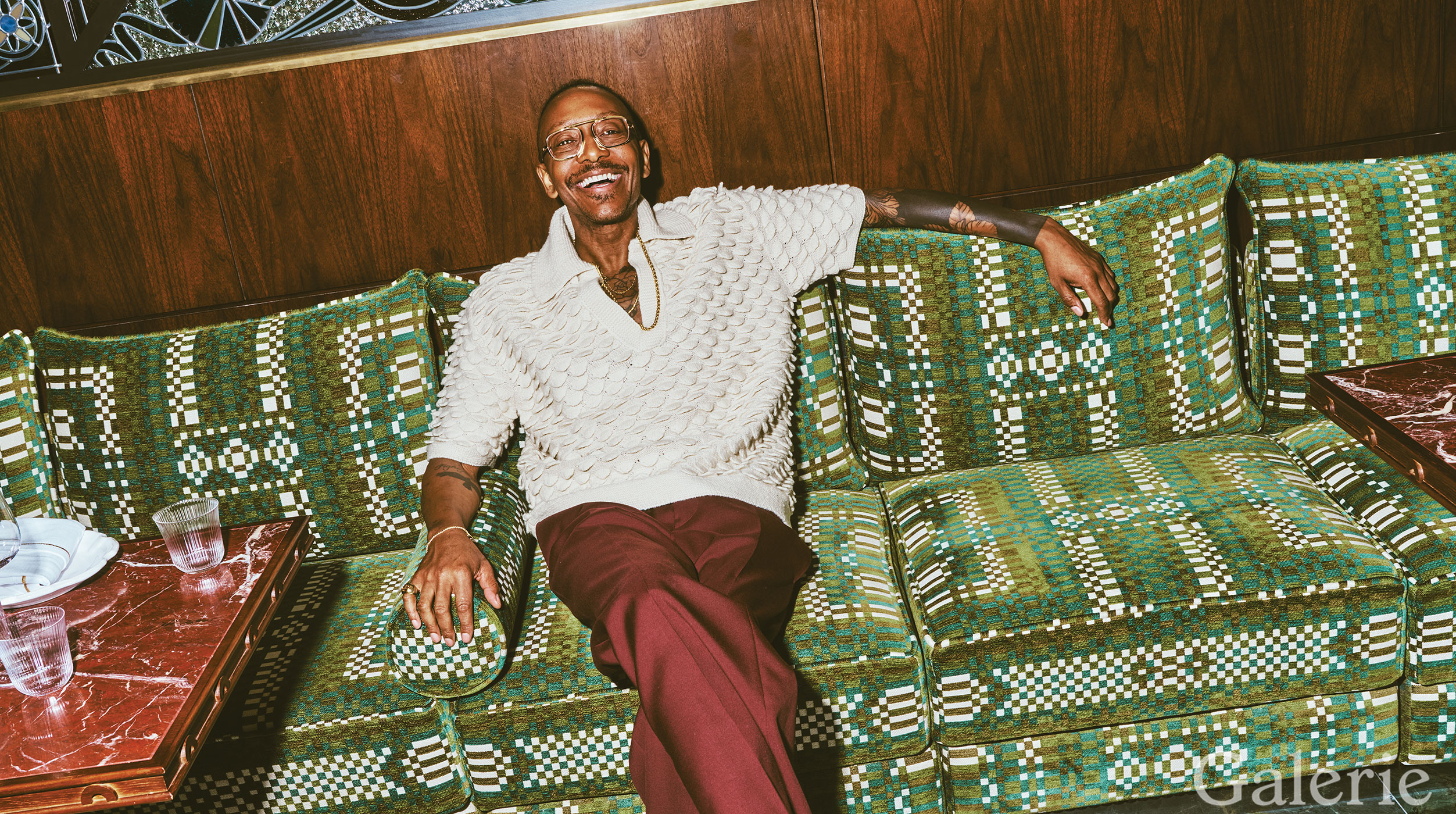 Smiling person in a sweater and glasses sitting on a patterned green couch in a cozy, wood-paneled room.