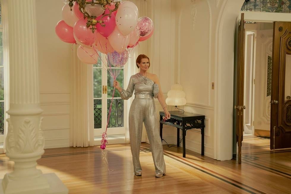 A woman in a silver jumpsuit holding pink and white balloons in an elegant room with wooden floors and ornate decor.